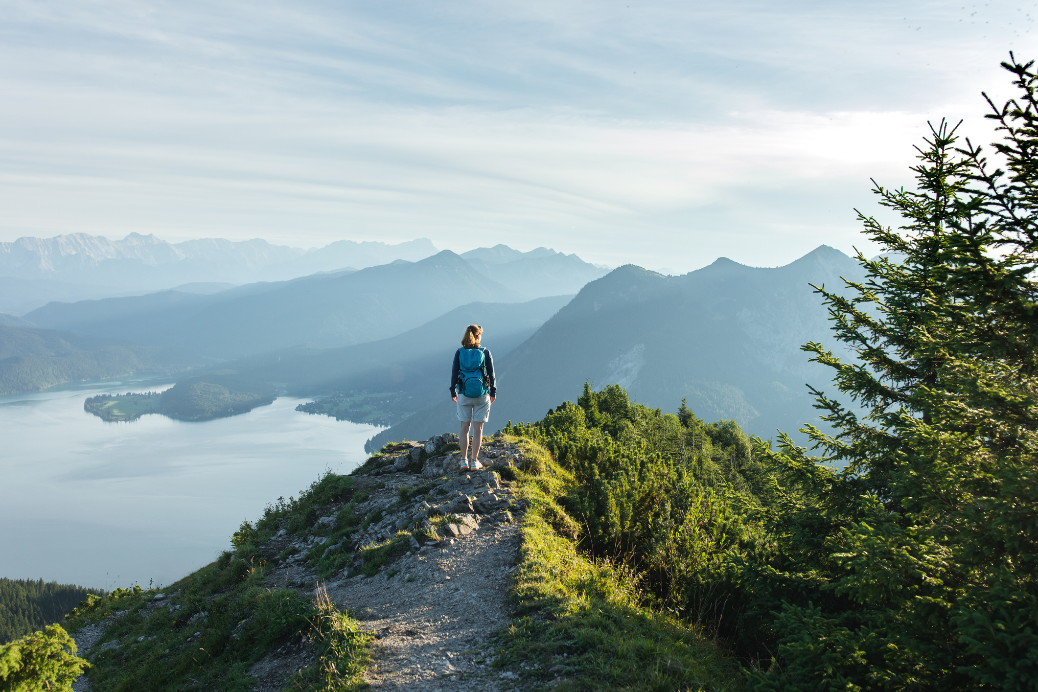 Woman climbing a mountain and standing at the top looking out onto a lake and mountains