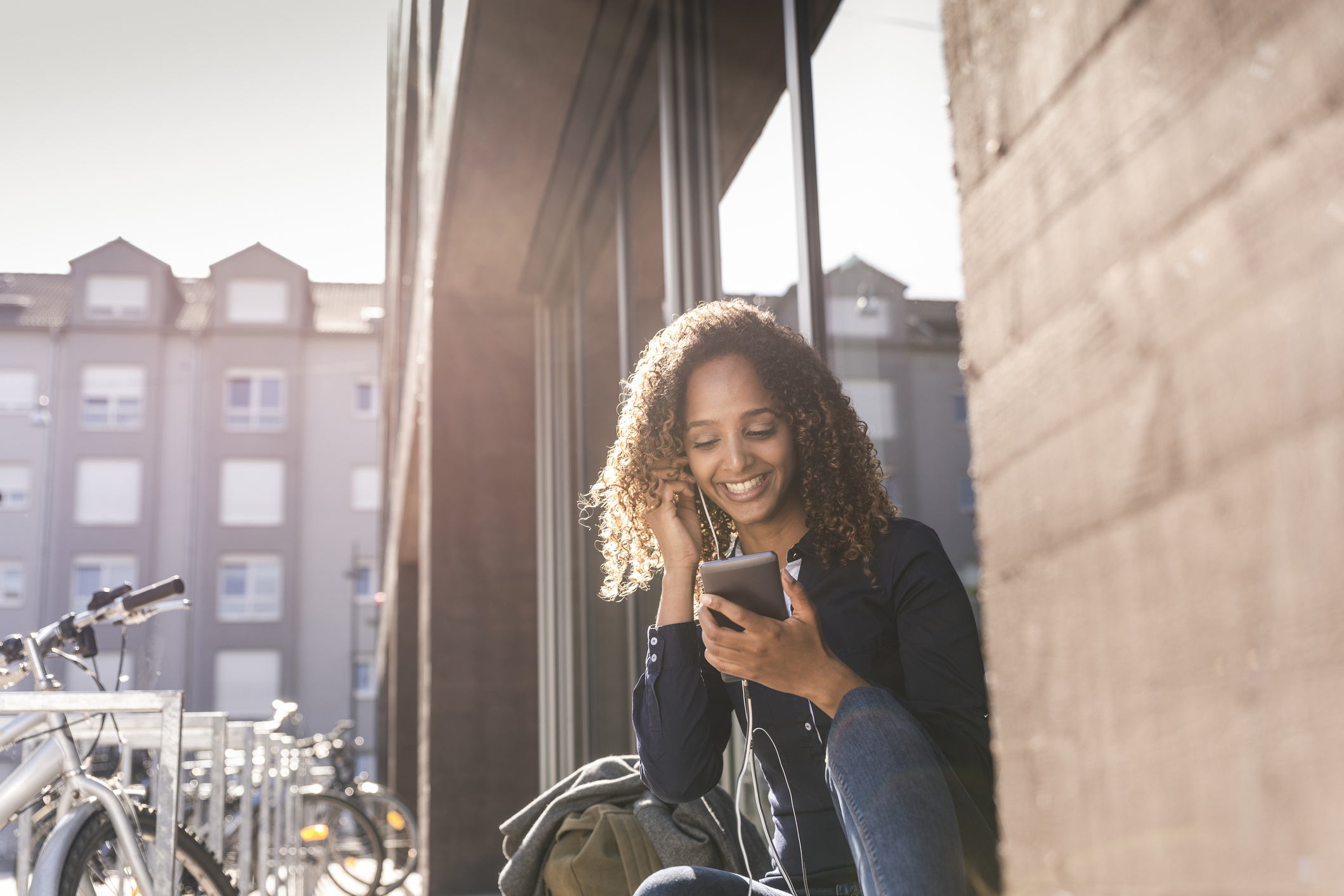 Young woman sitting in front of window in the city, using smartphone