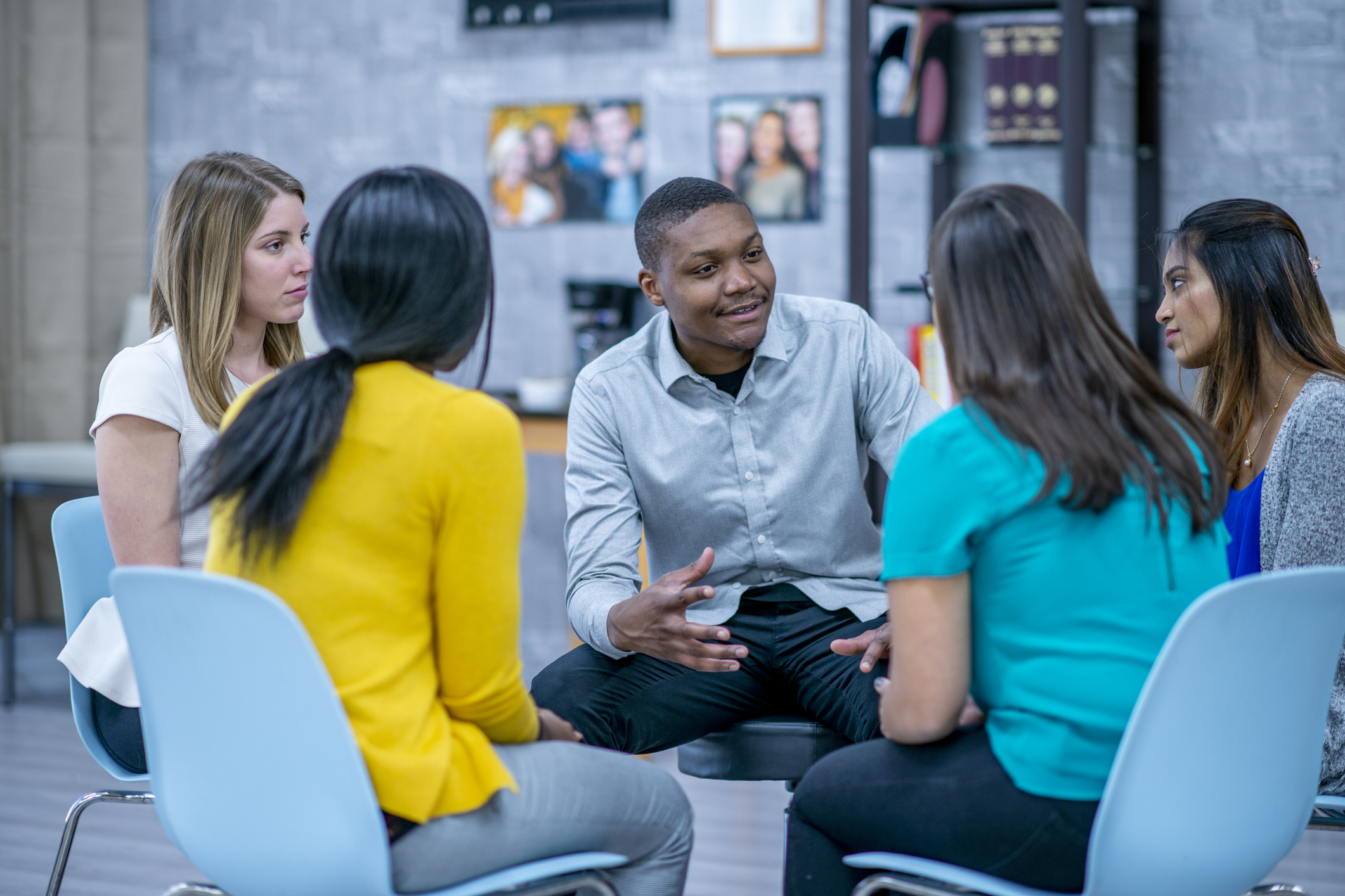 A diverse group of adults sit in a circle and listen to each other attentively. One man is speaking and smiling.