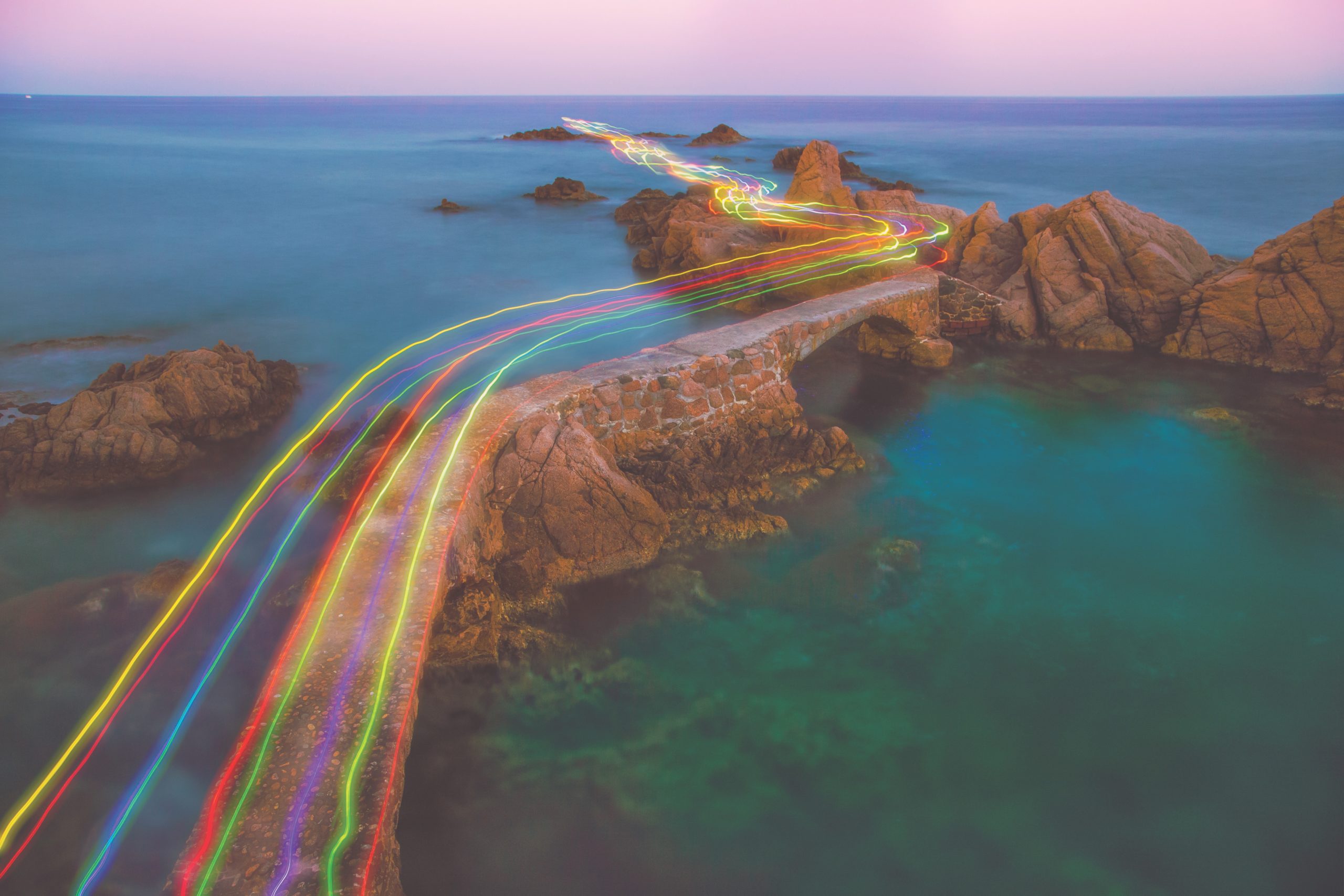 Long exposure of a colorful rainbow of light traces in a beautiful curved path between the rocks over the Mediterranean Sea in the Costa Brava shoreline on sunset.
