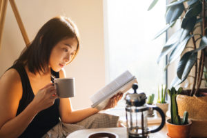 Beautiful young woman reading a book while drinking coffee, enjoying the morning sunlight.