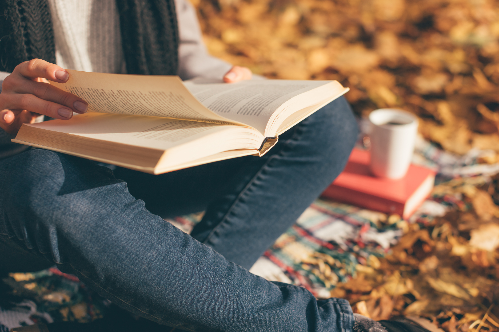 Cropped image of young woman sitting on blanket, reading book and drinking coffee or tea in autumn garden