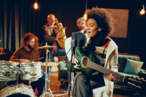 Mixed race woman singing and playing guitar while sitting on chair with legs crossed. In background drummer, saxophonist and bass guitarist.