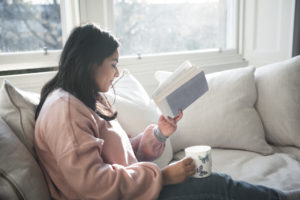 A British-Bangladeshi woman smiles as she reads a book and has a cup of tea while sitting on a white sofa next to a window in a flat in Edinburgh, Scotland, United Kingdom