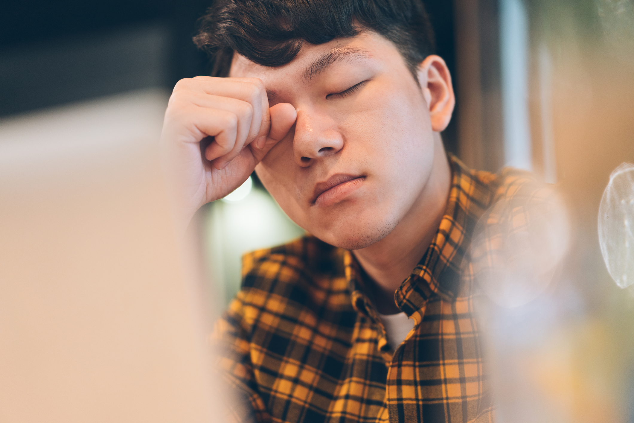 Exhausted young man rubbing eyes in cafe with laptop.