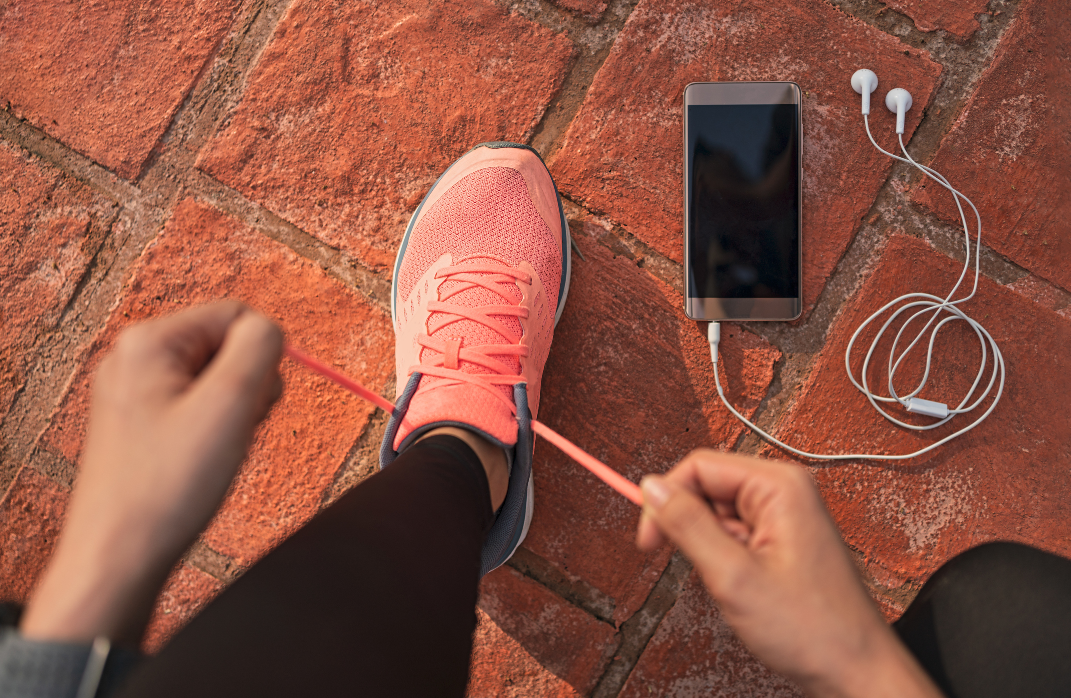 Runner woman tying running shoes laces getting ready for race on run track with smartphone and earphones for music listening on mobile phone. Athlete preparing for cardio training. Feet on ground.