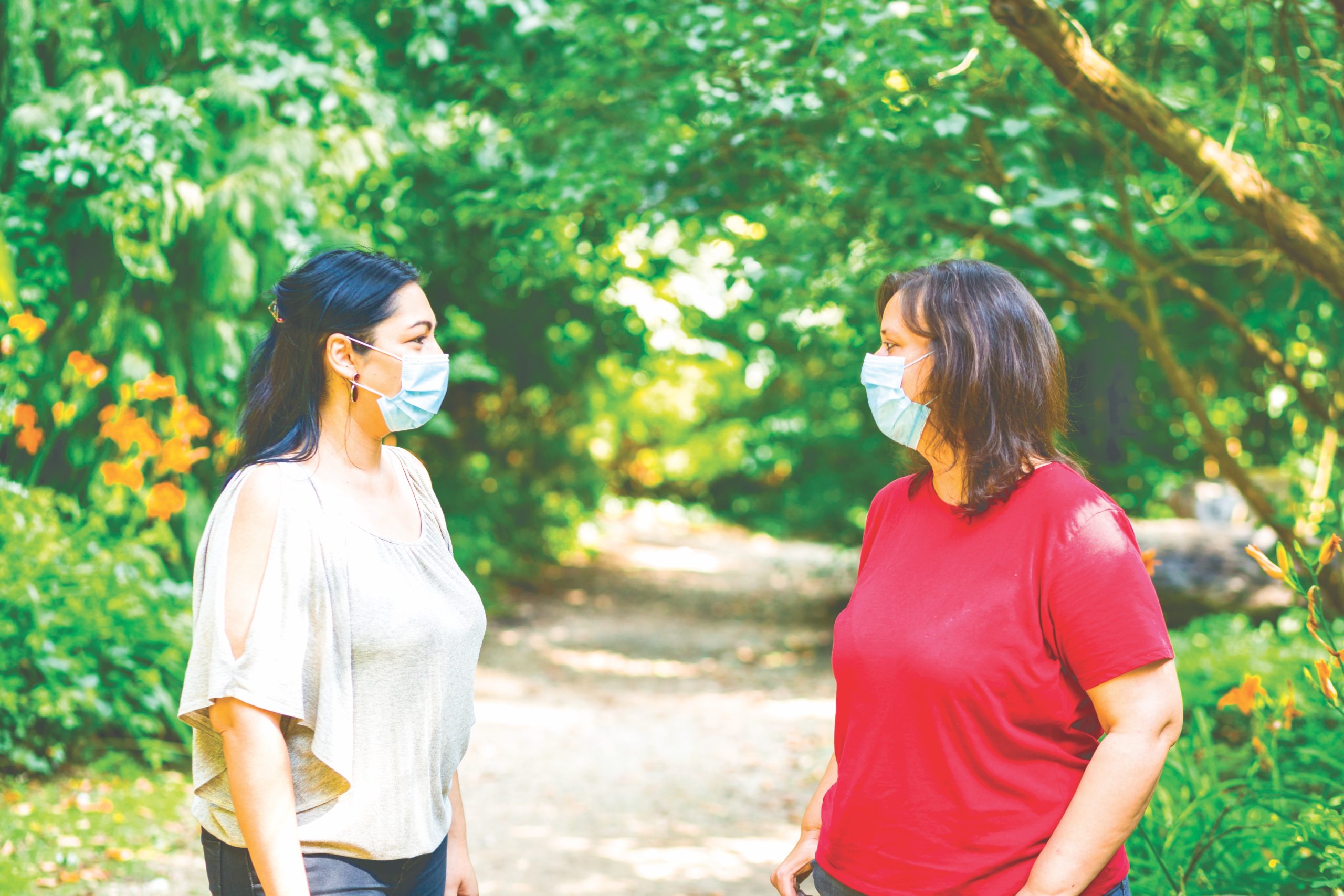 Close up color image depicting two friends, in their 30s walking in the park together while maintaining social distancing and wearing protective face masks during the Covid-19 pandemic.