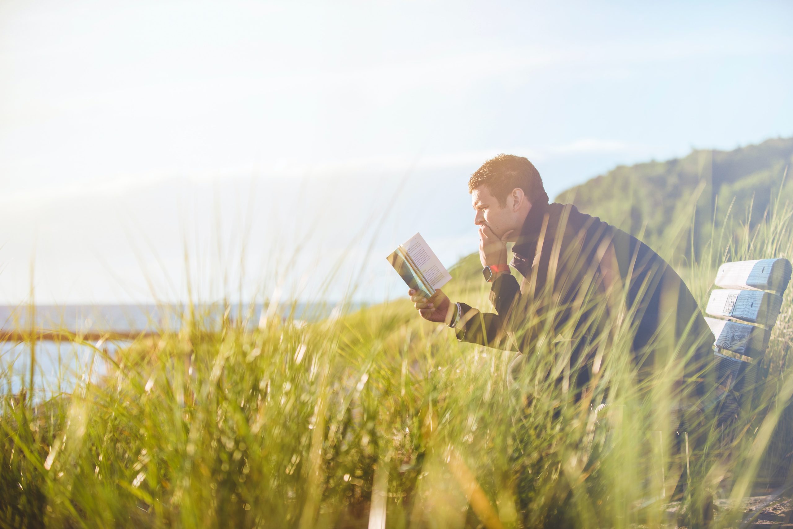 Person reading book near an ocean while sitting on a bench in the green grass.